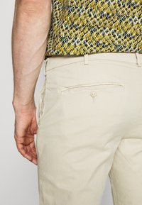 Beige bukser med en glat tekstur, der har en knappet baglomme og bæltestropper. Beklædningen er kombineret med en mønstret skjorte.