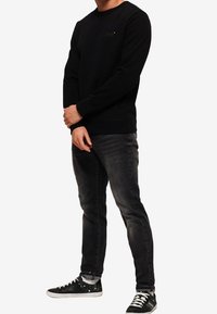 Felpa nera con polsini a coste, indossata sopra jeans scuri. Le sneaker presentano un design a trama. Uomo in piedi con le braccia incrociate.
