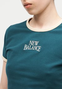 Κοντινό πλάνο προσώπου που φοράει μπλουζάκι New Balance σε τιρκουάζ χρώμα με εκρού γιακά και άκρες μανικιών, εμφανίζοντας το κεντημένο λογότυπο στο στήθος.