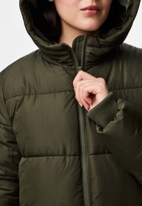 Casaco puffer verde-oliva com capuz, apresentando secções acolchoadas e um fecho frontal com zíper. As mangas são de malha, proporcionando um ajuste confortável.