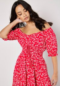 Rotes Blumenkleid mit kurzen Puffärmeln, gesticktem Taillenbund und einem Taillengurt mit Quasten. Das Muster umfasst weiße Blumen und Blätter.