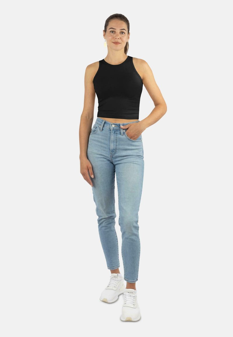 Frau in einem schwarzen ärmellosen Croptop, hochgeschnittenen hellblauen Jeans und weißen Sneakers, die mit einer Hand in der Tasche steht.