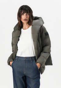 Olive green puffer jacket with a high collar, zip closure, and side pockets, worn over a white shirt and dark denim trousers.