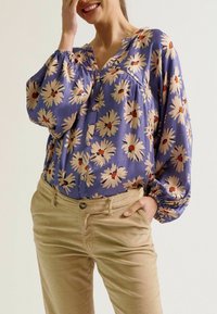 Blumenbluse mit langen Ärmeln, blauer Hintergrund, cremefarbene und rote Gänseblümchen, Knopfleiste, kombiniert mit hellbeige Cordhosen.