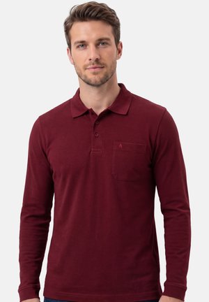 Młody mężczyzna w bordowej koszulce polo z długim rękawem i kieszenią na piersi, stojący na tle jasnoszarego tła.