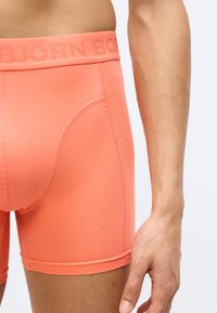 Κοραλλί μποξέρ με λεία, ελαστική ζώνη μέσης που φέρει ανάγλυφο όνομα μάρκας. Μαλακό ύφασμα, εφαρμοστό σχέδιο και κοντό μήκος στο πόδι.