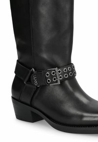 Bota de cuero negro hasta la rodilla con tacón cuadrado, que presenta una correa ancha adornada con ojetes y una hebilla como detalle. Textura suave.