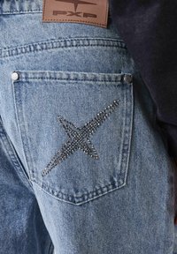 Jeans de mezclilla azul claro con un detalle cosido en forma de "X" adornado con piedras de strass en el bolsillo trasero. Etiqueta de cuero visible.