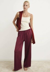 Top in raso bianco con spalline sottili, abbinato a pantaloni larghi bordeaux a gamba larga. Un cardigan rosso appoggiato su una spalla.