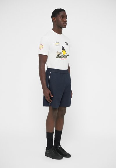 Λευκό T-shirt με γραφικό κείμενο και λεπτομέρειες, συνδυασμένο με σκούρο μπλε σορτς με πλευρικές ρίγες. Μαύρα αθλητικά παπούτσια ολοκληρώνουν την εμφάνιση.