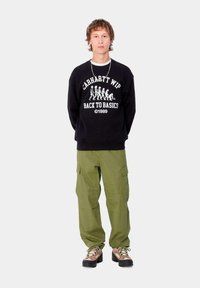 Sudadera negra con texto y gráficos en blanco, pantalones cargo verdes y zapatillas marrones con suelas negras; el modelo se encuentra de pie contra un fondo neutro.