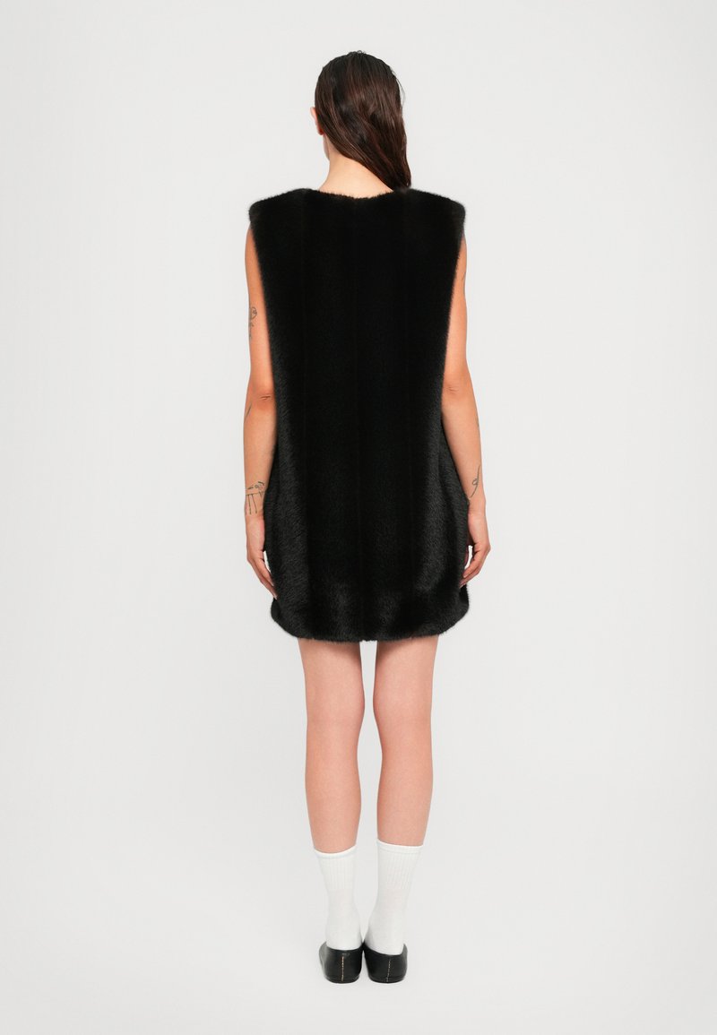 Woman with long dark hair wears a sleeveless black fur dress, white socks, and black shoes, standing facing a plain white background.