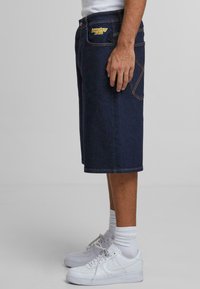 Shorts de mezclilla en azul oscuro con bordado amarillo. Cuentan con bolsillos y costuras en contraste. Combinados con zapatillas blancas y calcetines cortos.