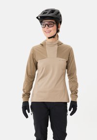 Maglia da ciclismo a maniche lunghe beige con cappuccio, caratterizzata da un pannello centrale in un beige scuro a contrasto. Abbinata a guanti neri e pantaloni.