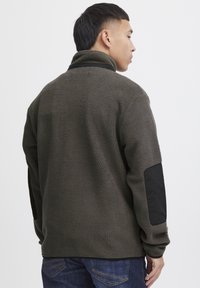Fleece pullover in donkergroen met een zachte textuur, hoge kraag en zwarte schouderpatches. Beschikt over lange mouwen en een relaxte pasvorm.