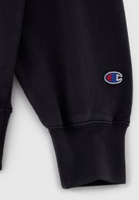 Sweatshirt noir avec des poignets et un ourlet côtelés. Présente un logo brodé en bleu et rouge sur la manche supérieure. Texture de tissu douce.