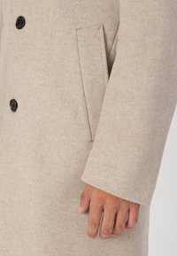 Abrigo de lana beige con una textura suave, featuring botones negros, un bolsillo de solapa, y mangas ajustadas. Primer plano de la manga y el área del bolsillo.
