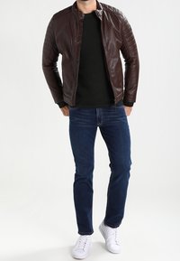 Veste en cuir marron avec épaules matelassées, pull noir, jean bleu et baskets blanches, debout devant un fond clair.