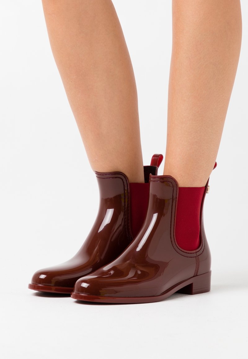 Botas de tobillo de goma brillantes en color marrón con paneles laterales elásticos rojos y tacón pequeño. Cuentan con una asa en la parte trasera para facilitar su uso.