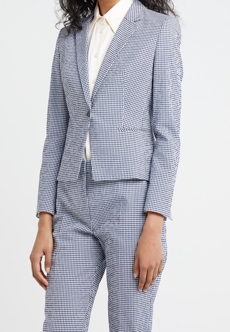 Femme portant un blazer à petits carreaux bleu et blanc et un pantalon assorti avec une chemise blanche boutonnée sur un fond uni.