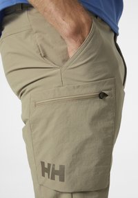 Beige cargobroek met een ritszak op de dij, met een gladde textuur en het "HH"-logo in een donkere schaduw.