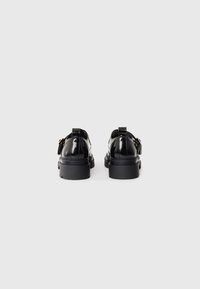 Black polished leather shoes with a chunky sole, featuring a secure buckle and decorative tassels, viewed from the back.