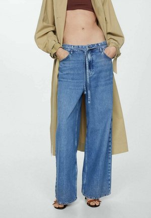 Osoba nosząca wysokie dżinsy o szerokich nogawkach z dopasowanym paskiem z denimu, brązowy krótki top, beżowy trencz oraz sandały na odkryte palce.