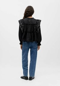 Blusa nera a maniche lunghe con bordi ondulati e gathered sulla schiena, abbinata a jeans blu chiaro e scarpe slip-on nere.