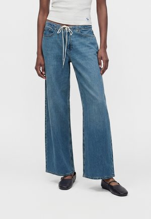 Άτομο που φοράει φαρδιά, χαλαρά μπλε τζιν με λευκό κορδόνι και μαύρα επίπεδα παπούτσια, στέκεται μπροστά σε ένα απλό φωτεινό φόντο.