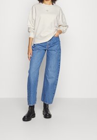 Felpa crema con polsini a costine, abbinata a jeans blu a vita alta. Il modello indossa robuste ankle boots neri. Design semplice e casual.