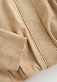 Tissu en daim beige avec des lignes de couture visibles et une manche pliée, mettant en valeur la texture et les détails des coutures.