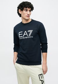 Temno modra jopica iz bombaža z veliko belim logotipom EA7 ter rebrastim okroglim izrezom. Dolgi rokavi in oblikovani manšeti.