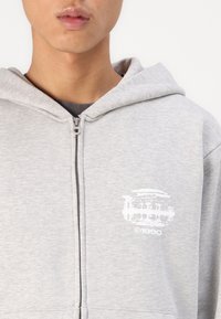 Haut à capuche zippé gris avec un logo à l'avant, fabriqué en tissu doux mélangé coton. Comprend une capuche avec cordon de serrage et un ourlet côtelé.