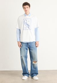 Balta īsām piedurknēm T-kreklā ar zilu "Calvin Klein" logotipu, uzvilkta uz gaiši zila pogājama krekla, savienota ar plēstiem gaiši ziliem džinsiem un baltām apaviem.