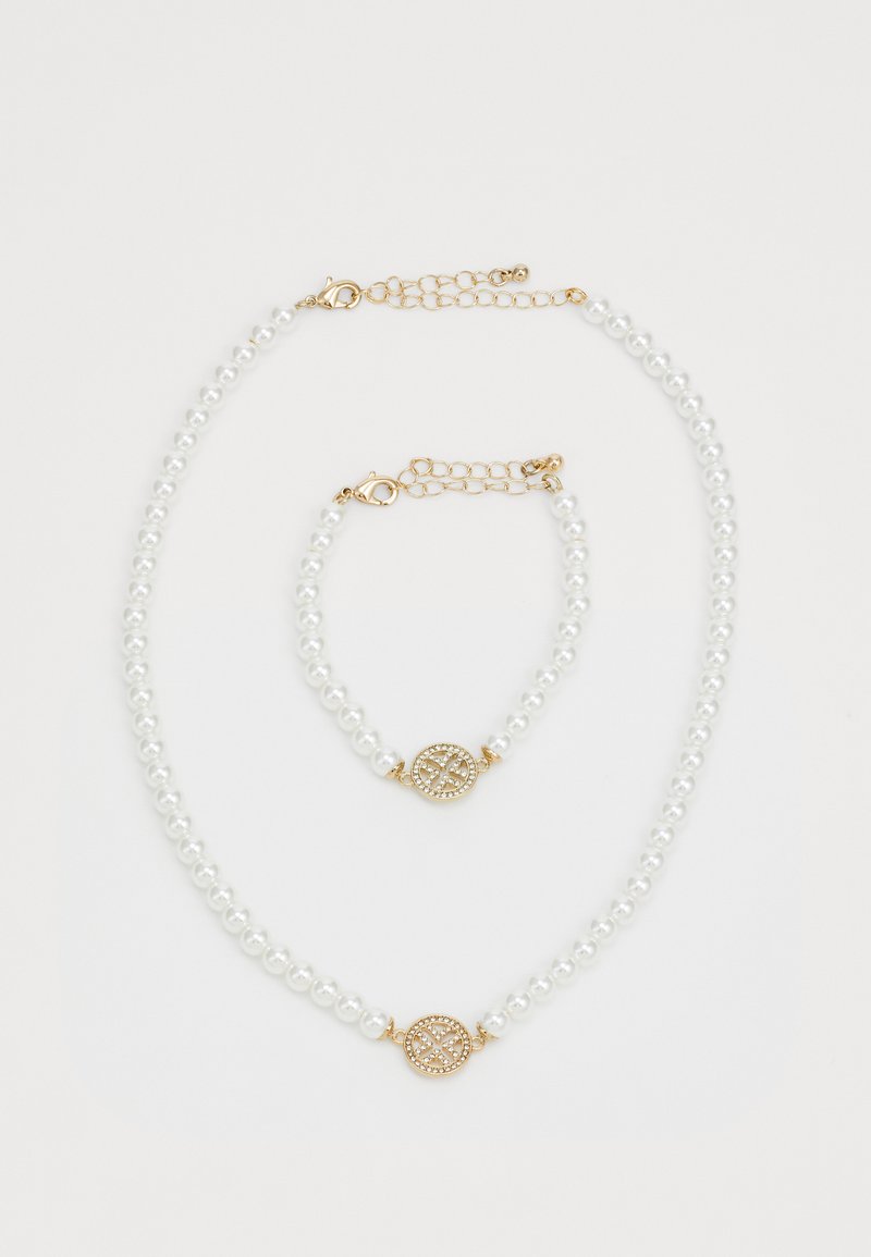 Parelketting en bijpassend armband met gouden sluitingen en ronde gouden bedel versierd met kleine kristallen op een witte achtergrond.