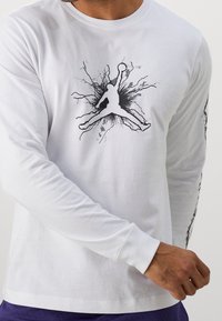 Valkoinen pitkähihaispaita, tehty puuvillasta, jossa musta Jordan-logo ympäröity räjähdyskuviolla. Hihassa on vastaava musta design-yksityiskohta.