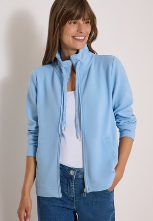 Kvinde iført lyseblå lynlåstrøje over hvid top og blå jeans, smilende og kigger til siden med den ene hånd i lommen.