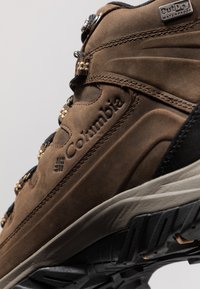 Columbia vattentät vandringskänga i brunt läder med texturerad yta, svarta detaljer och en robust svart gummisula, med spinngnaglar.