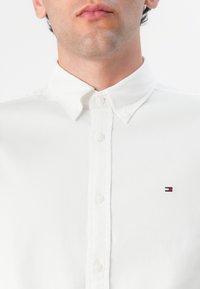 Camisa blanca de botones con un cuello estructurado, que presenta un pequeño logotipo bordado en el pecho. Tejido texturizado con un acabado suave.