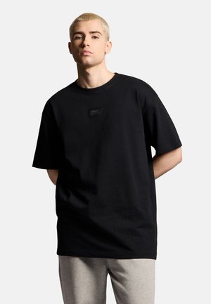 Młoda osoba z krótkimi blond włosami, ubrana w oversize czarny T-shirt i jasnoszare spodnie na jednolitym tle.