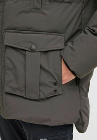 Chaqueta acolchada gris con un gran bolsillo frontal, cierre de botones a presión, bolsillos laterales para las manos y puños acanalados. Tejido suave con un brillo sutil.