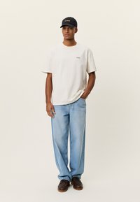 Νεαρός άνδρας που φοράει κρεμ μπλουζάκι, ανοιχτό μπλε τζιν, σκούρα καφετιά παπούτσια και μαύρο καπέλο, στέκεται με το ένα χέρι στην τσέπη του.