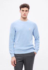 Világoskék kötött pulóver texturált anyaggal, bordázott mandzsettával és szegéllyel, bal oldalon a mellkason egy kis kör alakú logóval. Szürke nadrággal párosítva.