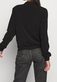 Sort bomberjakke med glat stof, ribstrikkede manchetter og linning. Løs pasform med faldede skuldre, stylet med mørke denimjeans.