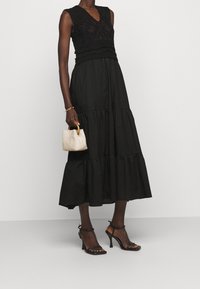 Frau in einem ärmellosen schwarzen Schichtenkleid, schwarzen High-Heelsandalen, goldener Armbanduhr und hält eine kleine beigefarbene Handtasche mit Holzgriffen.