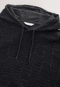 Svart luvtröja med strukturerat tyg med geometriska mönster. Inkluderar dragsnören och en etikett med varumärket "NEXT."