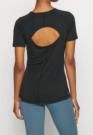 Personne portant un top noir à manches courtes avec une grande découpe ovale dans le haut du dos et un pantalon bleu ajusté.