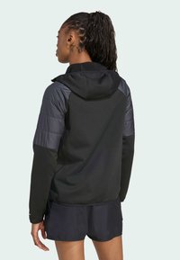 Žena s pletenicom nosi crnu kapu i kratke hlače, okrenuta leđima prema svijetloj pozadini, ističući svoju sportsku odjeću.