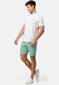 Cămașă polo albă cu mâneci scurte, având un logo mic, asortată cu pantaloni scurți verde mentă și adidași albi, stând într-o poziție relaxată.