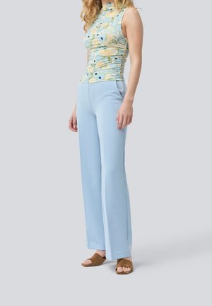 Femme portant un haut fleuri sans manches, un pantalon large bleu clair et des sandales ouvertes marron, debout devant un fond uni.